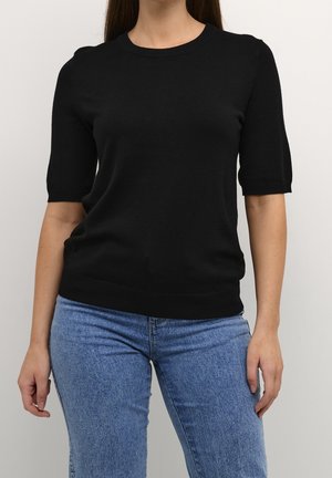 Femme portant un haut en maille noir à manches courtes associé à un jean taille haute bleu clair sur un fond uni.