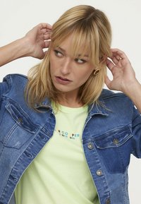 Jeansjacke mit klassischem Schnitt, blauer Farbe und Metallknöpfen. Trägt eine hellgrüne T-Shirt mit buntem Text.