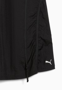 Veste noire en tissu léger, avec un design froncé sur le côté, une fermeture éclair, un ourlet à cordon de serrage et un logo Puma blanc dans la partie inférieure.
