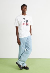 Vit kortärmad T-shirt med svart och röd grafisk tryck, ihop med ljusblå baggy jeans och svarta mönstrade skor. Grön golvbakgrund.