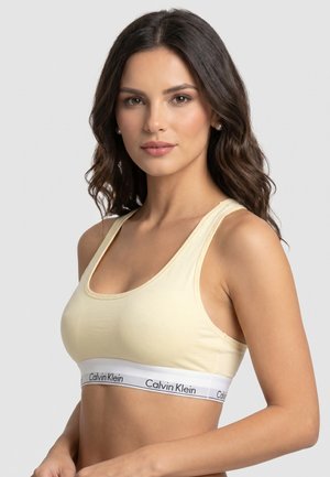 Jonge vrouw met lang, donker golvend haar draagt een lichtgele Calvin Klein sportbeha, kijkt iets naar rechts op een effen grijze achtergrond.