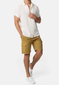 Camisa blanca de manga corta con botones en tela texturizada combinada con pantalones cortos cargados color caqui con bolsillos laterales, usada con zapatos blancos sin cordones.