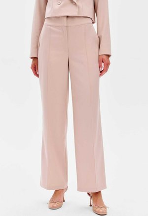 Pantalons larges beiges sur mesure en tissu lisse, dotés de coutures avant et de poches latérales. Assortis à des talons beiges clairs avec un accent en forme de nœud.