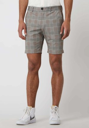 Homme portant un short gris à carreaux et des baskets blanches montantes, debout devant un fond clair uni.