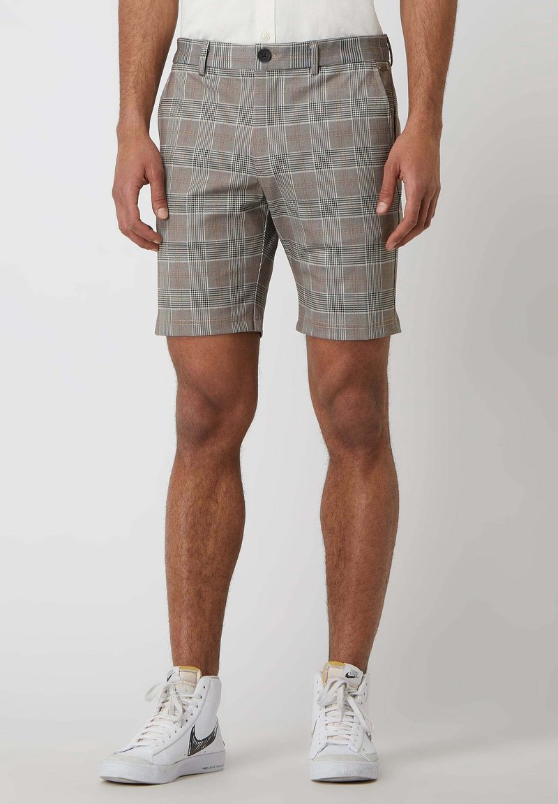 Homme portant un short gris à carreaux et des baskets blanches montantes, debout devant un fond clair uni.