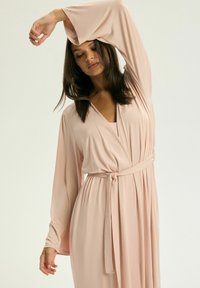 Jasnoróżowa, długosleeve sukienka kopertowa z głębokim dekoltem w kształcie litery V, pasek do wiązania w talii, lekko opadający materiał i miękka, gładka tekstura.