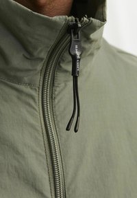 Šviesiai žalia zip-up striukė su aukštu apykakle, kurios juodas užtrauktukas ir virvės reguliavimas. Lygus pavirčius su minimaliu matomų siūlių kiekiu.