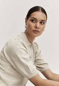 Beige gestrickter Pullover mit einem Polo-Kragen, kurzen puffartigen Ärmeln und dekorativen Knöpfen, getragen mit goldenen Creolen. Glatte Textur.