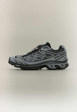 Zapatilla de trail running Salomon en negro y gris con patrones geométricos y sistema de cordones rápidos, mostrada de lado sobre un fondo neutro.