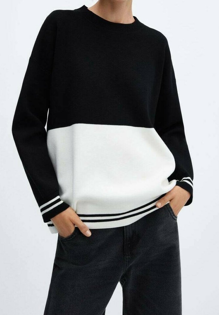 Personne portant un pull bicolore noir et blanc avec des poignets et un ourlet rayés, mains dans les poches, associé à un jean foncé.