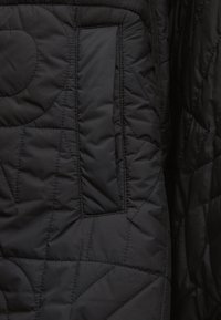 Μαύρο καπιτονέ ύφασμα με ραμμένα γεωμετρικά σχέδια και κάθετη ορθογώνια τσέπη στην αριστερή πλευρά.