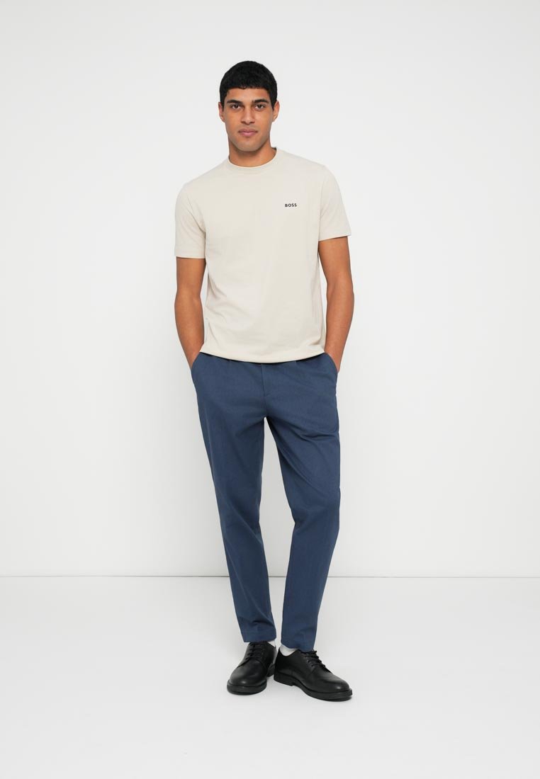 T-shirt beige à manches courtes avec le logo "BOSS", associé à un pantalon bleu marine et des chaussures noires. Le mannequin est debout devant un fond blanc uni.