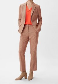 Blazer e pantaloni sartoriali in un motivo a scacchi marrone chiaro, abbinati a un top a V di colore arancione brillante. Stilizzati con stivaletti beige alla caviglia.