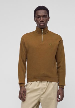 Homme portant un pull marron à fermeture éclair quart de zip et un pantalon beige, debout devant un fond clair uni.