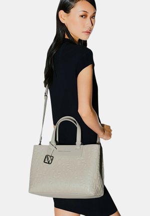 Donna in abito nero che porta una borsa beige chiaro con scritte in rilievo e un ciondolo logo in metallo sulla spalla.