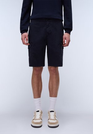 Pantalones cortos de carga en color marino, confeccionados en una tela liviana, con dos bolsillos laterales y cierre de botón. Combinados con zapatillas beige y calcetines blancos.