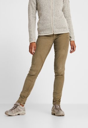 Dolna część ciała osoby ubranej w jasnobrązowe spodnie sztruksowe, szarą kurtkę na zamek i beżowe buty trekkingowe, stojącej z lekko rozstawionymi nogami.