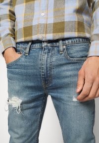 Jeans de mezclilla azul claro con una rodilla deshilachada, que presentan un diseño clásico de cinco bolsillos y costuras amarillas. Llevados con una camisa de cuadros.