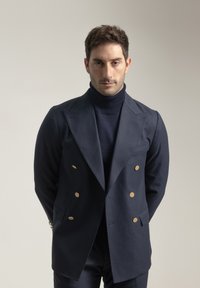 Blazer azul marino de doble botonadura con botones dorados, diseño ajustado y solapas con muesca. Debajo, se ve un cuello de tortuga azul marino a juego.