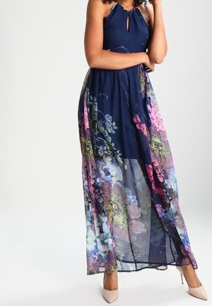 Femme portant une robe maxi bleu marine sans manches avec un voile transparent à motif floral et des escarpins beiges pointus, posant les bras croisés.