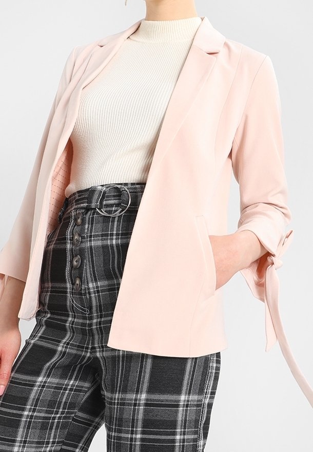 Blazer rose clair à la coupe cintrée, avec un col à revers et des manches retroussées. Associé à un pantalon à carreaux gris taille haute et un haut côtelé.