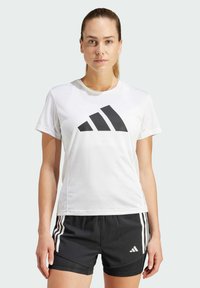 Weißes Sport-T-Shirt mit kurzen Ärmeln und einem großen schwarzen Adidas-Logo, kombiniert mit schwarzen Shorts, die weiße Streifen und Logo-Highlights aufweisen.
