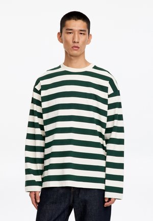 Jeune homme portant une chemise à manches longues à rayures horizontales vertes et blanches et un jean foncé, debout sur un fond blanc uni.