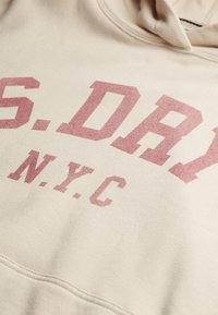 Beżowa bluza z kapturem z dużym, wyblakłym różowym nadrukiem z napisem "S.DRY" i "N.Y.C." w wyraźnej czcionce. Miękka tkanina z kieszenią typu kangur.