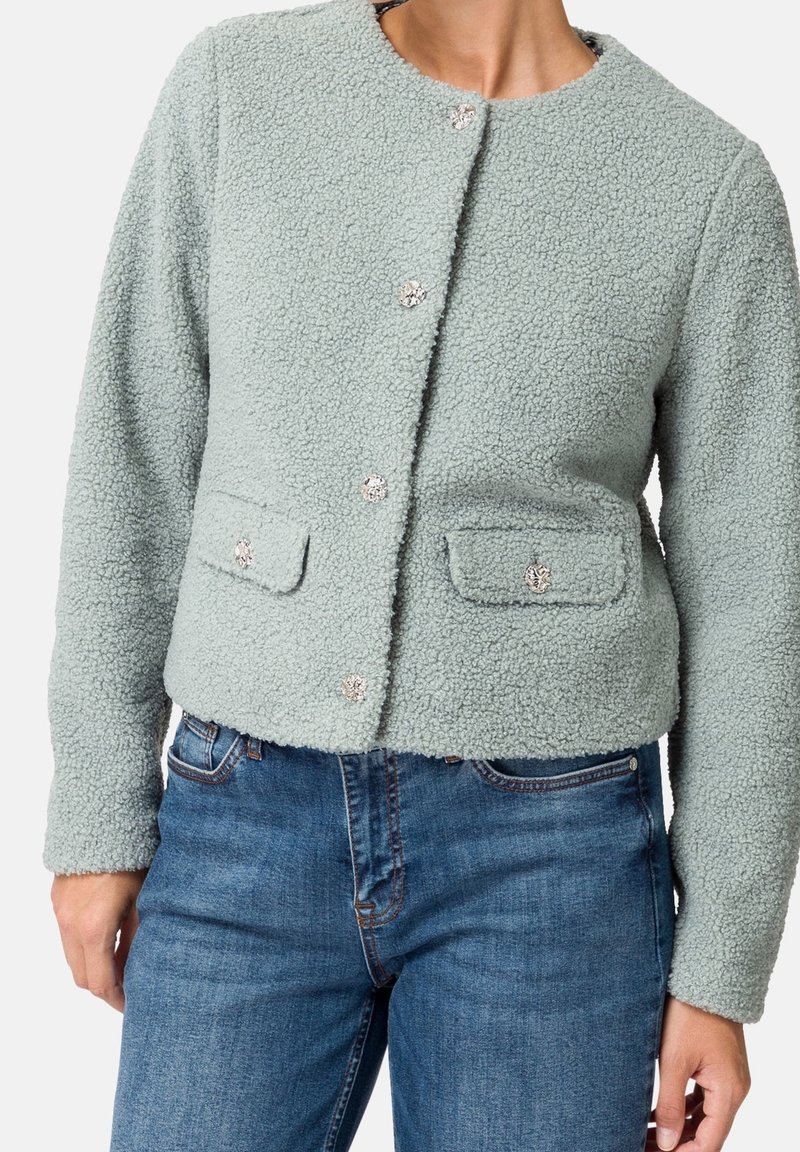 Femme portant une veste boutonnée texturée vert clair avec poches à rabat, associée à un jean en denim bleu, debout devant un fond blanc.