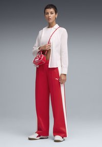 Red track pants with white side stripes, a white button-up shirt, and a small red patent handbag. White sneakers with a tan sole.