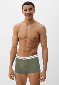 Smiling fit young man with short curly hair wearing olive green boxer briefs with a white waistband, standing against a plain light background.