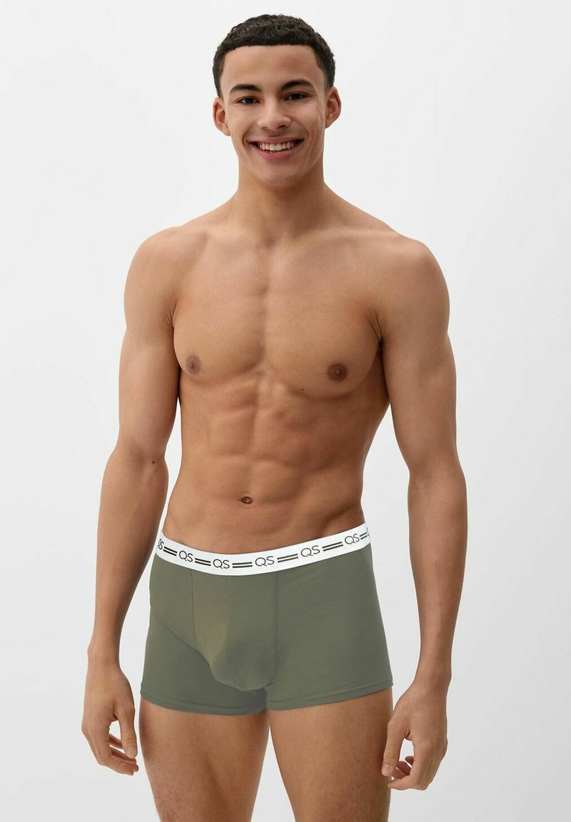 Smiling fit young man with short curly hair wearing olive green boxer briefs with a white waistband, standing against a plain light background.
