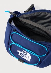 Námořnicky modrá látková taška s jasně modrými doplňky a logem. Má zipové zapínání a vnitřní kapsu, texturovaný materiál a pevnou konstrukci.