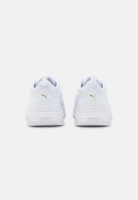 Baskets de sport blanches avec une tige en mesh texturé, accents de logo dorés, col rembourré et semelles en caoutchouc. Vue arrière montrant la boucle au talon.