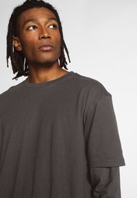 Tumši pelēks īsām piedurknēm T-krekls ar apaļu kakla izgriezumu, ar mīkstu tekstūru un brīvu piegriezumu. Nav redzamu rakstu vai logo.