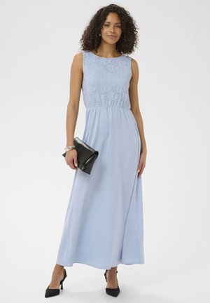 Femme portant une robe maxi bleu clair sans manches avec un corsage en dentelle, tenant une pochette noire et portant des talons noirs ainsi qu'un bracelet argenté.