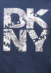 Λευκά γράμματα τύπου block "DKNY" γεμάτα με ασπρόμαυρη εκτύπωση εφημερίδας σε σκουρό μπλε ύφασμα.