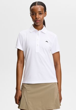 Donna che indossa una polo bianca a maniche corte con un piccolo logo e una gonna beige, in piedi davanti a uno sfondo semplice e chiaro.