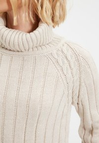Woman wearing a cream-colored, ribbed knit turtleneck sweater with cable knit details on the shoulder and upper arm.