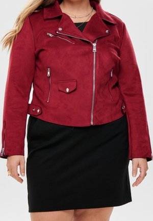 Frau trägt eine bordeauxrote Bikerjacke aus Kunstwildleder mit silbernen Reißverschlüssen und einem schwarzen knielangen Rock.