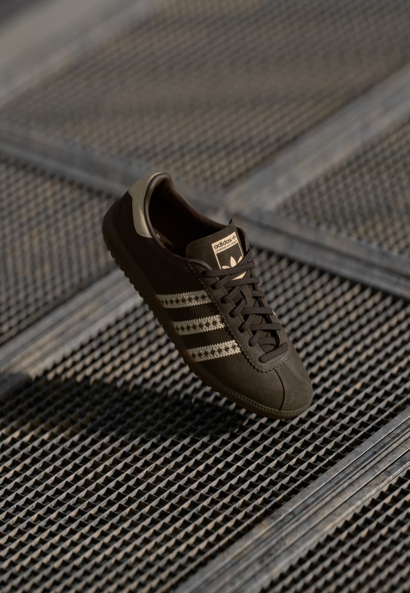 Sneaker in suede marrone con tre strisce dorate, suola in gomma e punta cucita. Presenta chiusura con lacci e logo del marchio sulla linguetta.
