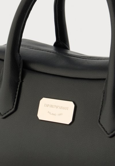 Μαύρη δερμάτινη τσάντα χειρός με δύο λαβές και χρυσή πινακίδα με την επιγραφή "Emporio Armani Milano 1981" μπροστά.