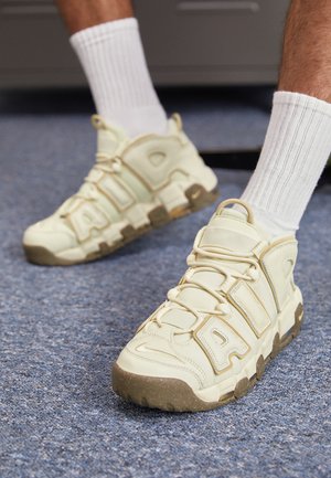 Stopy w beżowo-brązowych butach sportowych Nike Air z białymi prążkowanymi skarpetkami na niebieskim wykładzinowym podłożu, pokazane z niskiego kąta.