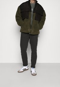 Groene fleecejas met zwarte bovenkant van stof, twee borstzakken, grijs shirt eronder, zwarte broek en witte sneakers met zwarte strepen.
