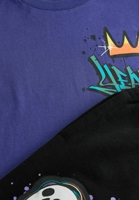Violet t-paita, jossa on graffitityylisiä grafiikoita, kuten kruunu ja roiskeefekti, kerrostettuna mustan kankaan vaatteelle, jossa on pääkalloasu.