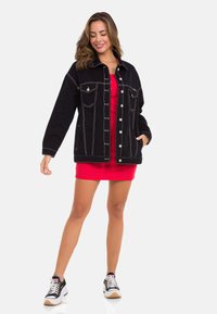 Schwarze Denimjacke mit kontrastierender Naht, zwei Brusttaschen und Knopfverschluss, kombiniert mit einem roten, figurbetonten Kleid und schwarzen Sneakers.