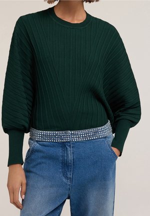 Ciemnozielony prążkowany sweter z bufkami, zestawiony z niebieskimi dżinsami i srebrnym paskiem z ćwiekami, na osobie stojącej z jedną ręką w kieszeni.