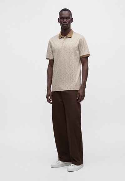 Homme debout portant un polo beige à motif, un pantalon large marron foncé et des baskets blanches sur un fond clair uni.