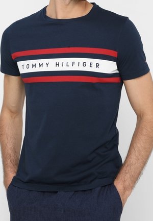 Mand iført navyblå T-shirt med korte ærmer, røde og hvide vandrette striber samt teksten "TOMMY HILFIGER" tværs over brystet.