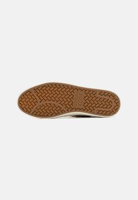Hallitusseentest valmistatud spordijalatsite välispind, millel on pruun herringbone muster ja valge äär. Jalanõul on "Converse" bränding.